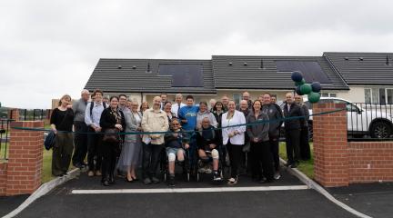 Westfield Housing Association opening event for 6 Bungalows. Colleagues from WHA, residents, and Cumberland Council are smiling at the camera in front of the new development, behind an opening ribbon and balloons. 