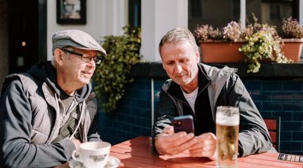 Two men sat outside at pub one with beer, one coffee, look at mobile phone