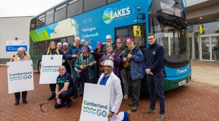 A group of people pose in front of a blue double‑decker bus branded “The Lakes Connection,” with destinations including Workington and Cockermouth displayed on the side. Several individuals are holding musical instruments, such as ukuleles. Others hold large white signs that read “Cumberland GO”. 