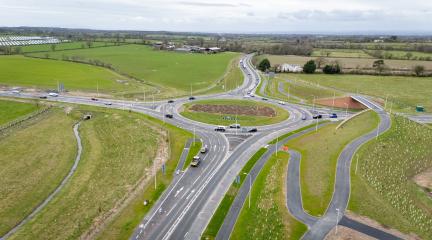 Aerial view of a newly constructed roundabout connecting multiple roads, surrounded by green fields and landscaped verges. Several vehicles are travelling through the junction, with footpaths, cycle routes and road markings clearly visible across the site.