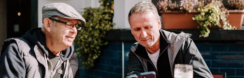 Two men sat outside at pub one with beer, one coffee, look at mobile phone