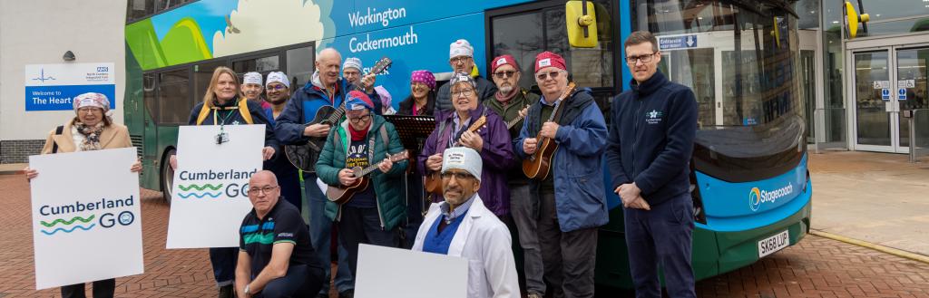 A group of people pose in front of a blue double‑decker bus branded “The Lakes Connection,” with destinations including Workington and Cockermouth displayed on the side. Several individuals are holding musical instruments, such as ukuleles. Others hold large white signs that read “Cumberland GO”. 
