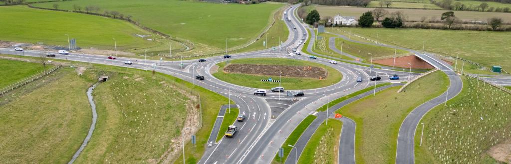 Aerial view of a newly constructed roundabout connecting multiple roads, surrounded by green fields and landscaped verges. Several vehicles are travelling through the junction, with footpaths, cycle routes and road markings clearly visible across the site.